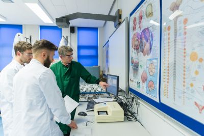 Two students wearing white coats in the lab, watching a member of staff wearing a green lab coat gesture towards a desktop computer