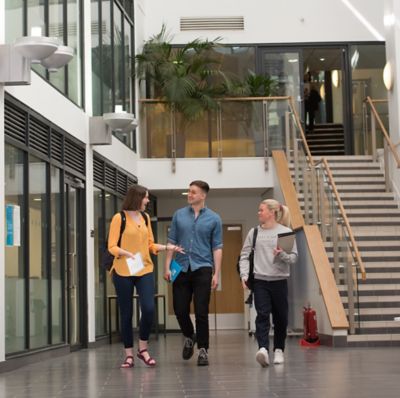 Postgraduate students Christopher Lanyon (British); Hayley Mills - blonde (British) and Heather Collis red shirt (British) walking in the Mathematical sciences building atrium. May 2019 by Simon Litherland.