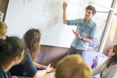 A physics lecturer writing on a whiteboard, presenting to a group of students