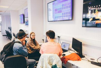 Three undergraduate students huddled around laptops in the product design studio underneath screens portraying student work