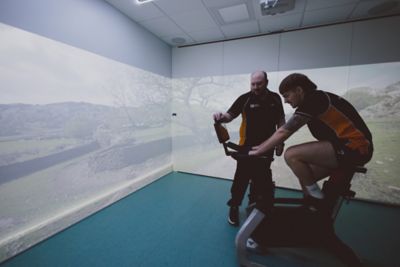 Assistant Professor watching an undergraduate student on a Wattbike, in the Immersive Suite