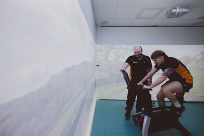 Assistant Professor watching an undergraduate student on a Wattbike, in the Immersive Suite