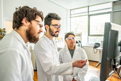 Postgraduate students Tom Burwell, Hiba Azim and Ethan Norman, wearing lab coats and safety goggles looking at a computer monitor showing cyclic voltamogram