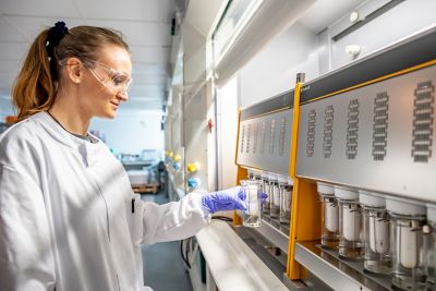 Senior Technician holding a glass flask containing a food sample in the analytical lab wearing safety goggles.