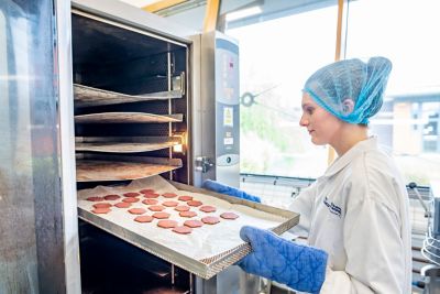 Undergraduate student Katherine Tolson taking baked products from an oven wearing lab coat, hair net and oven gloves