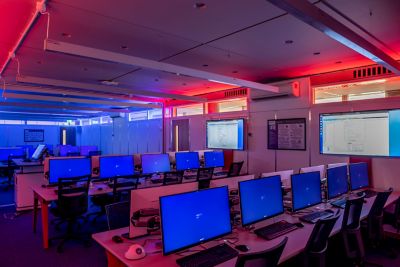 Cyber security lab full of desks with computer and monitors switched on, room lit red and blue